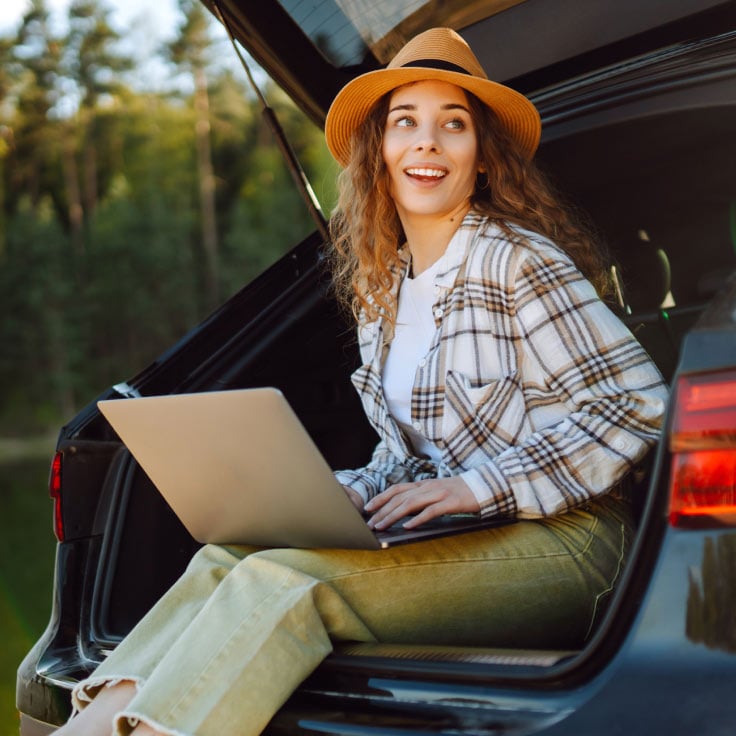 A young woman views her laptop while siting in the back of an SUV that's parked in a wooded environment.